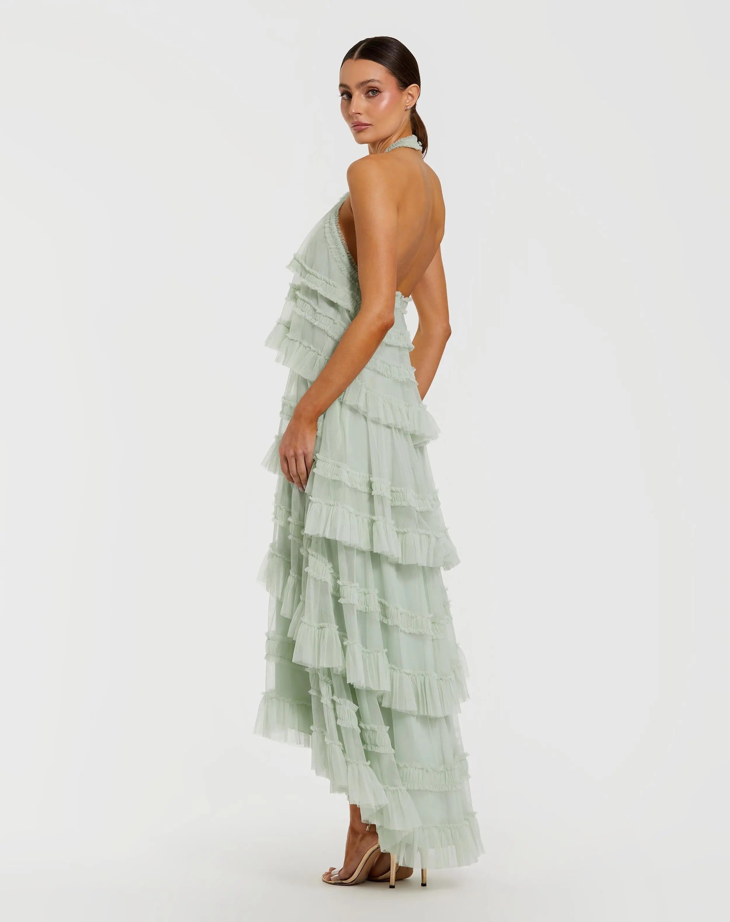 A woman poses sideways against a plain white background, showcasing the elegant tiered layers and high-low hemline of the Mac Duggal 8276 Tea-Length Party Dress in pale green, styled with nude high heels.